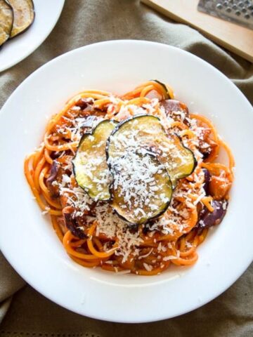 Pasta alla norma in a white plate, garnished with fried eggplant circles and grated ricotta salata cheese.