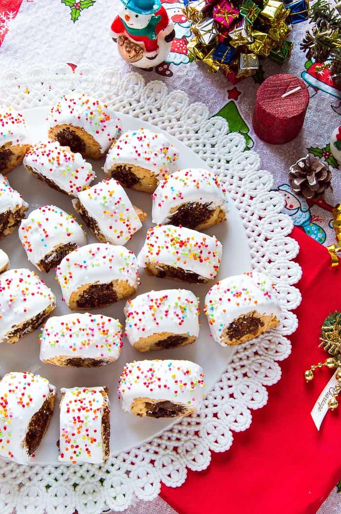 Cuccidati cookies covered with icing and decorated with sugar sprinkles with Christmas decorations in the background.