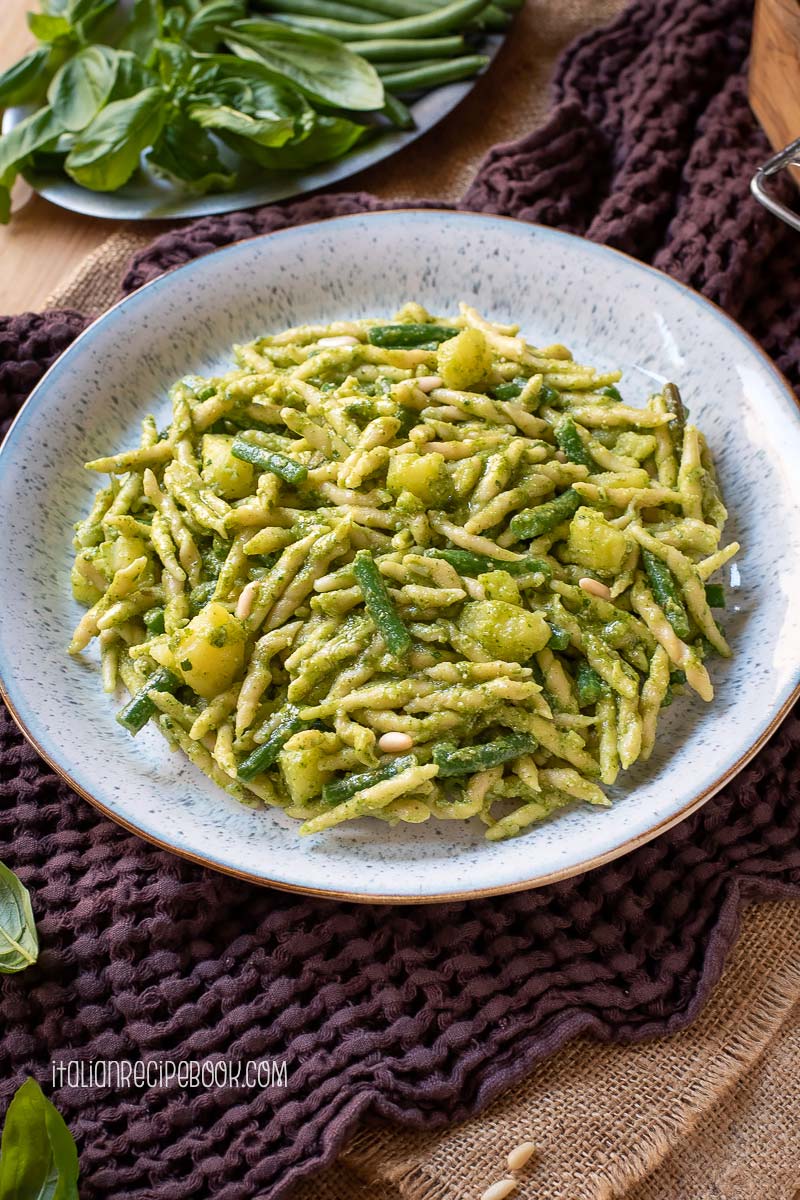 Trofie pasta with pesto, potatoes and green beans on a plate with basil leaves in the background.