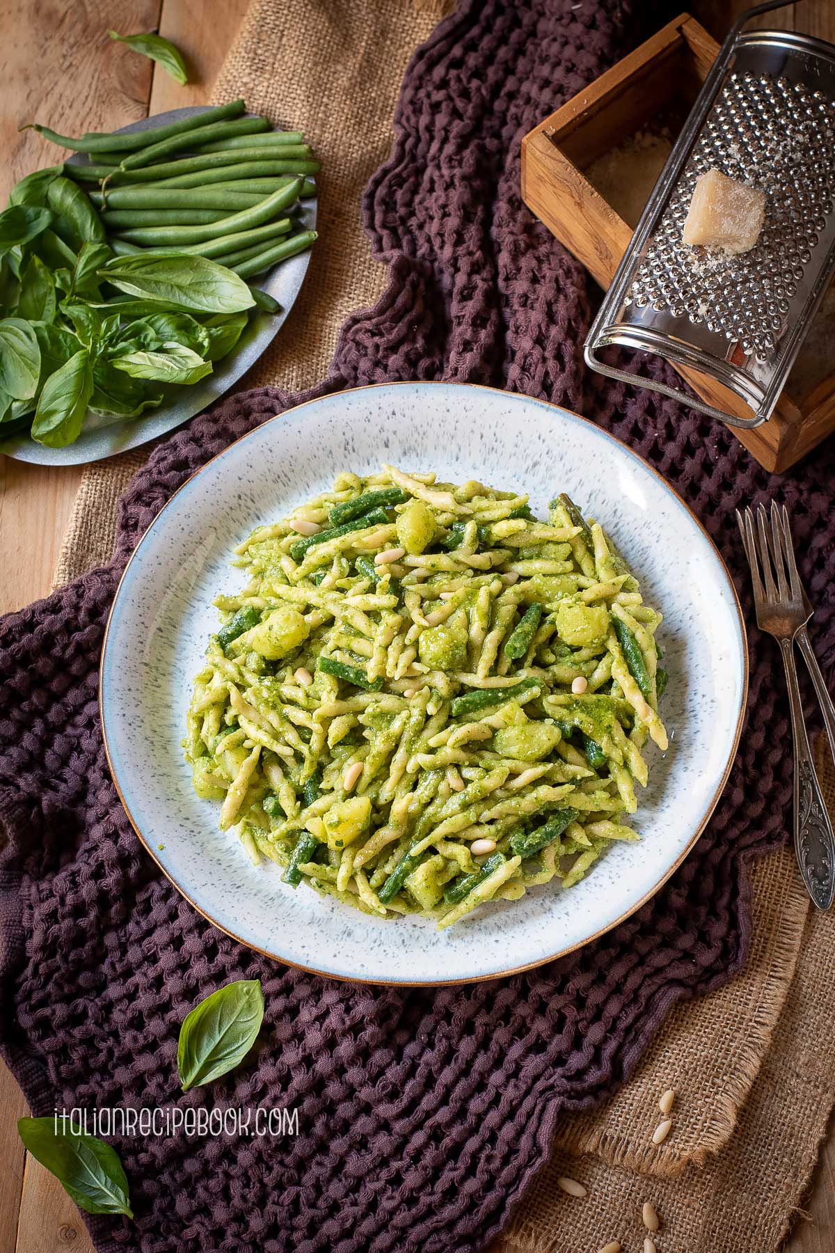 Pasta al pesto on a place with Parmesan cheese grater, fresh basil leaves and green beans laid in the background,