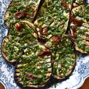 Grilled eggplant steaks topped with parsley olive gremolata and sun-dried tomatoes on a blue and white platter.