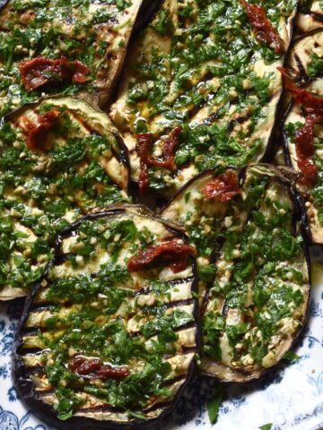 Grilled eggplant steaks topped with parsley olive gremolata and sun-dried tomatoes on a blue and white platter.