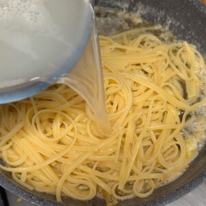 Pouring starchy pasta cooking water into the lemon butter and linguine in a pan.