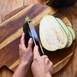 Slicing a whole eggplant lengthwise into steaks on a wooden board with a chef's knife.